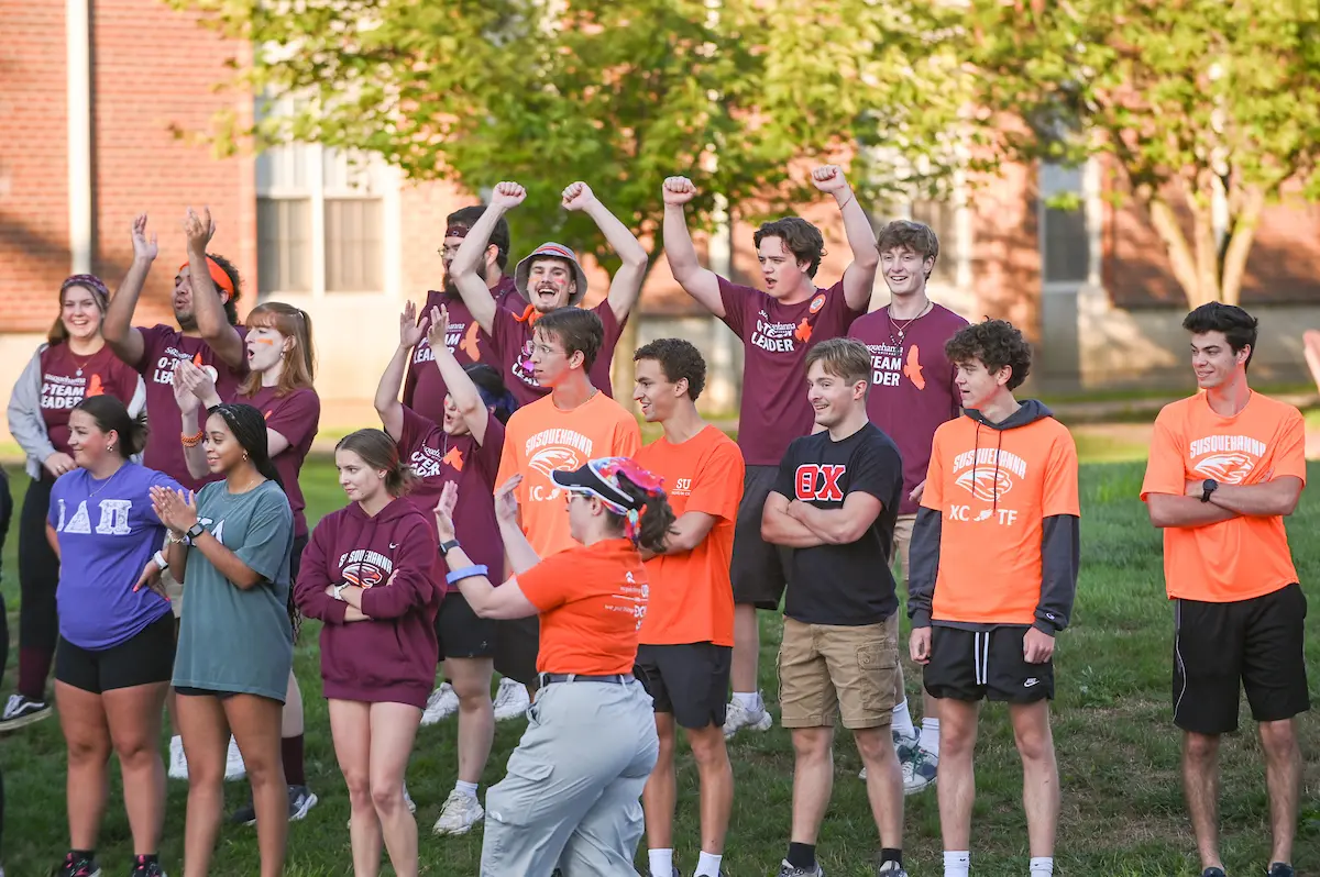 A group of college students stands outdoors in colorful shirts, cheering and clapping. Some wear maroon, others orange or green. Trees and a brick building are in the background. The scene is all about energetic, joyful campus spirit.