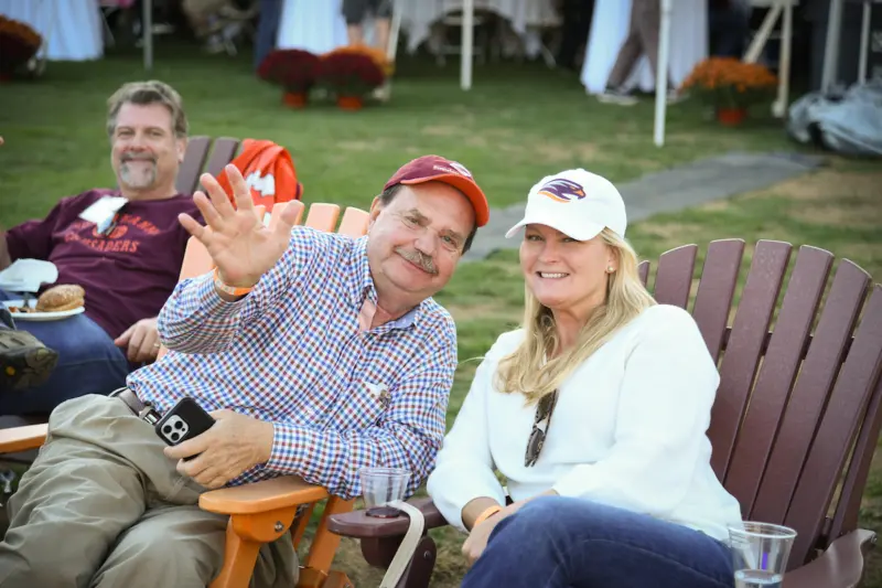 A man and a woman sit outdoors on wooden chairs during a homecoming reunion. The man, in a red cap and checkered shirt, waves at the camera while the woman, wearing a white cap and sweater, smiles warmly. Another man sits in the background with tables and plants behind them.