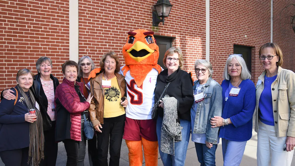 A group of women attending an event poses with a mascot in a bird costume wearing a white jersey, outside against a brick wall. Smiling and dressed in casual attire, they appear to be enjoying the moment together.