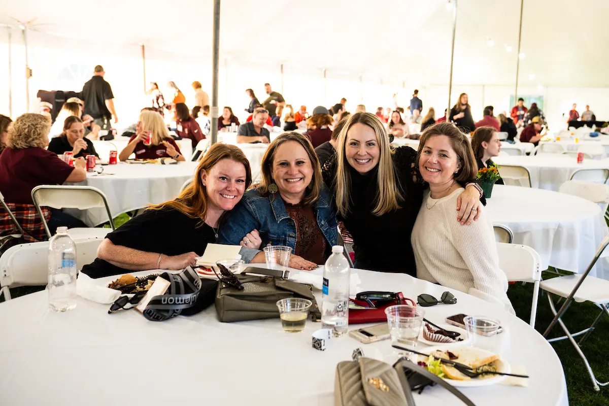 Four alumni women, smiling and posing together at a round table inside a large event tent. The table is adorned with drinks and personal items. In the background, more attendees are seated at other tables, adding to the lively atmosphere.