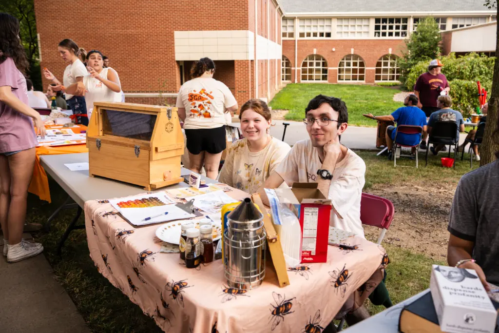 Two people smile at a table adorned with honey-related items and bee-themed decorations, showcasing sustainability initiatives. A bee-patterned tablecloth adds charm as they sit outdoors next to a brick building, with others nearby and more tables in the background.