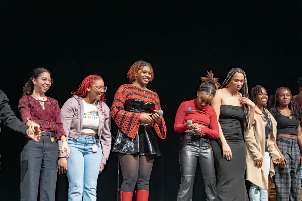 A group of people stands on stage smiling and holding microphones, showcasing their colorful, stylish outfits against a black background. It's a perfect example of 