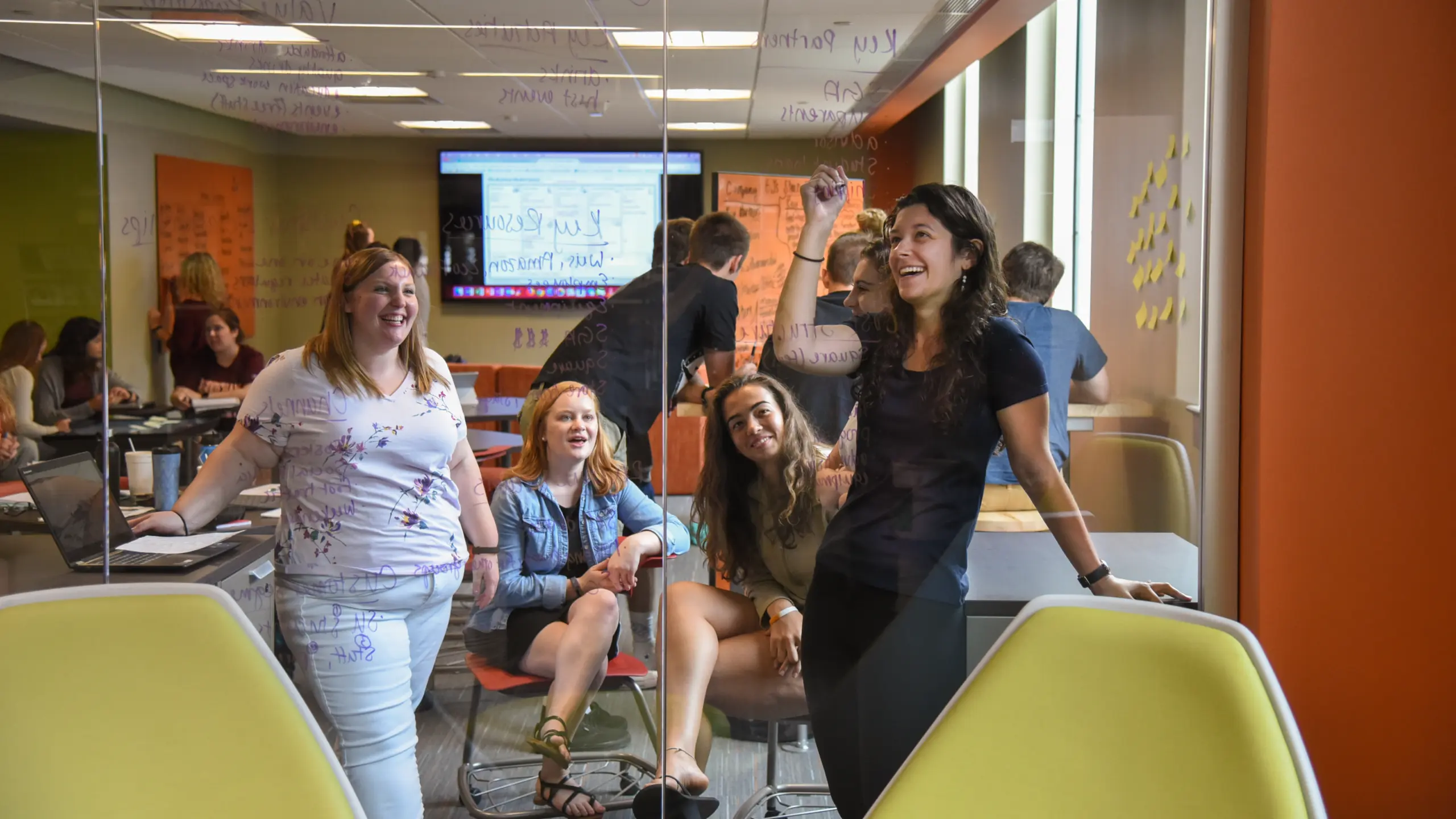 A group of people in a modern office setting, some sitting, some standing. They are engaged in discussion, with notes on a clear glass board in the background. Laptops and papers are on the tables, creating a collaborative atmosphere.