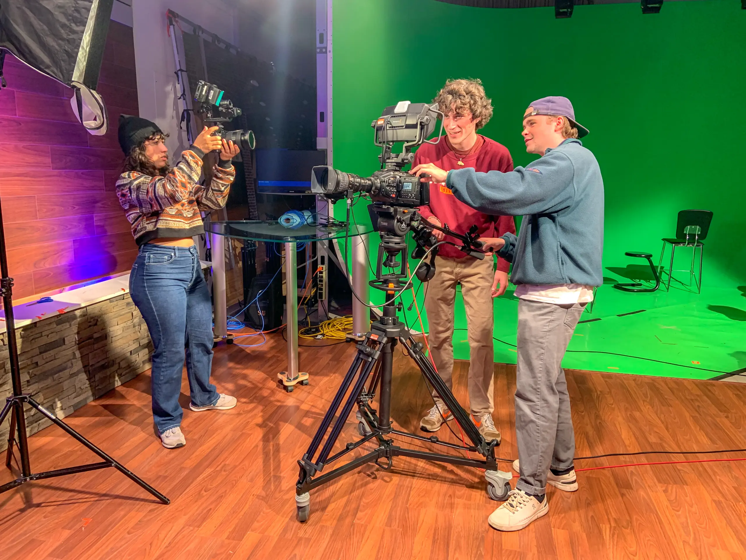 Three people in a studio film setting. Two are adjusting a large camera on a tripod, while another person films them. The background has a green screen and studio lights, with wooden flooring and a modern decor.