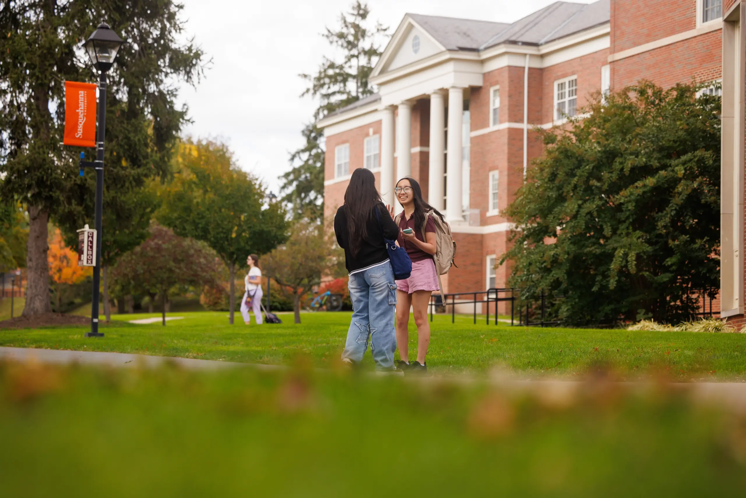 Two people are talking on a grassy area near a large brick building with columns. One wears a black jacket and jeans, the other wears a pink shirt and skirt. Trees and a lamppost are in the background, with others walking nearby.