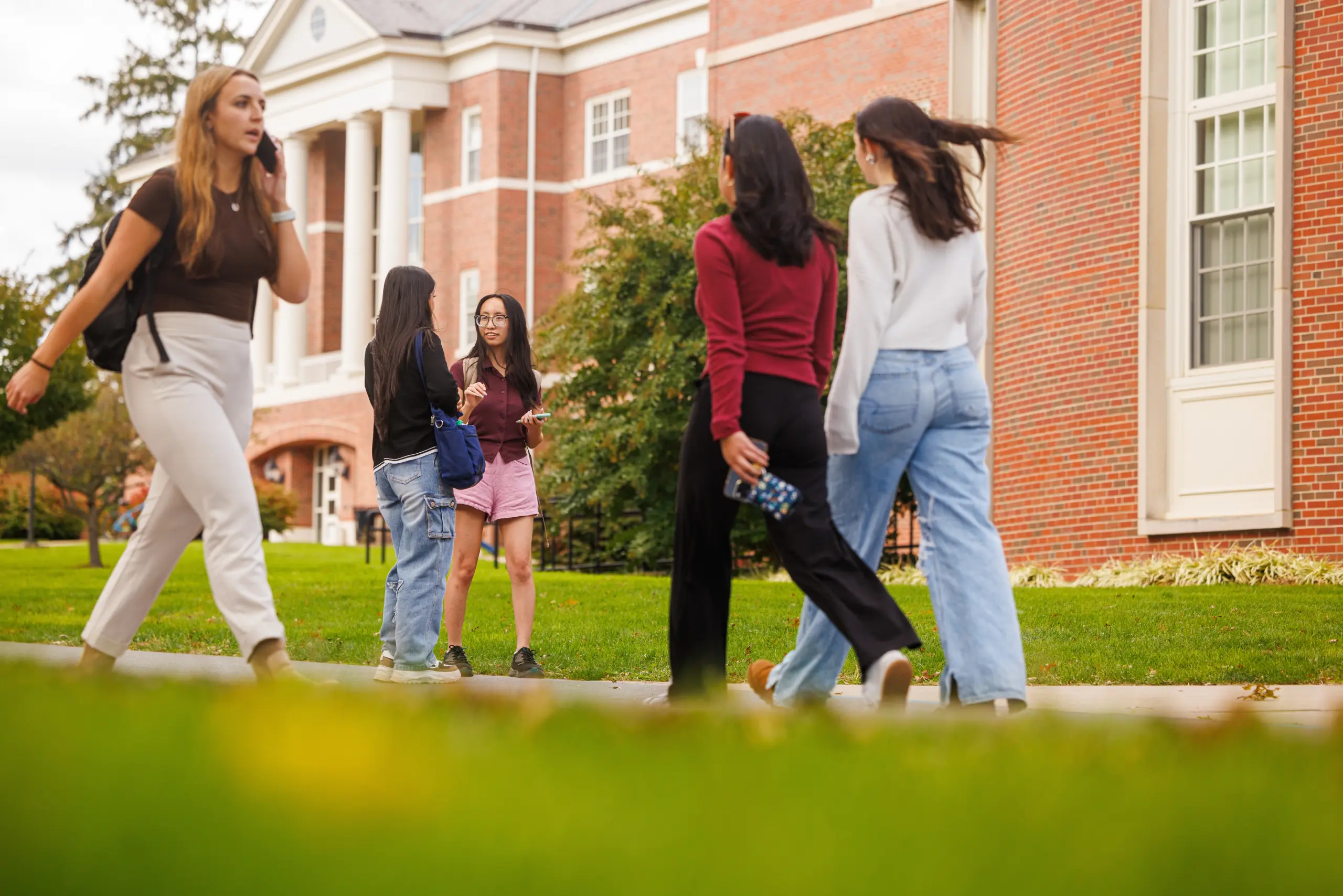 A group of people walk and converse on a college campus. They are surrounded by brick buildings and green lawns. Some are dressed casually, and one holds a phone. The scene is lively and vibrant.