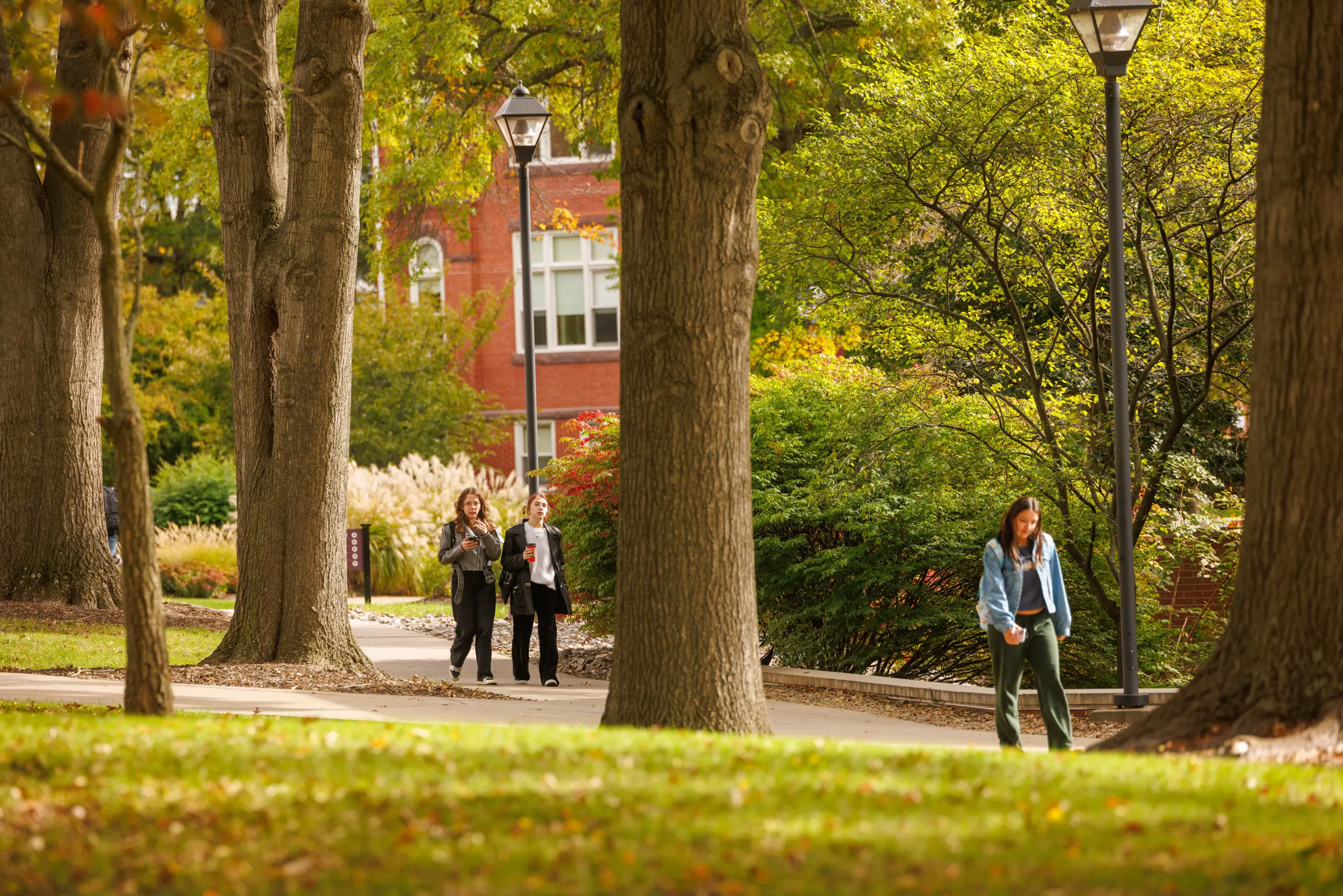 A woman walks through a tree-lined path on a sunny day. In the background, two people walk together carrying papers. A red brick building and green foliage are visible. Streetlamps dot the landscape.