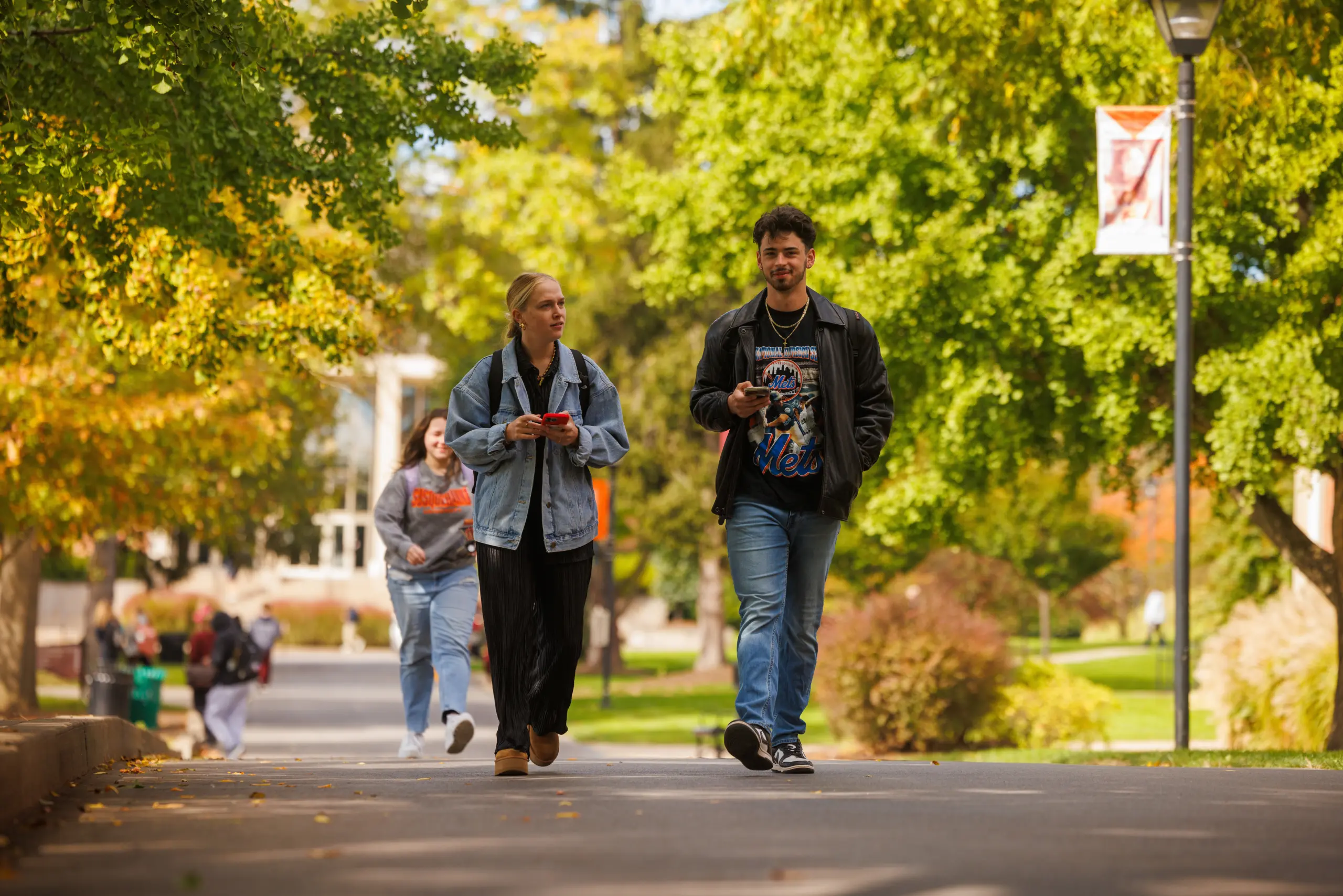 Two people walk together on a tree-lined path, with a woman in a denim jacket and a man in a black jacket. Another person follows in the background. The surroundings are lush with green trees and dappled sunlight.