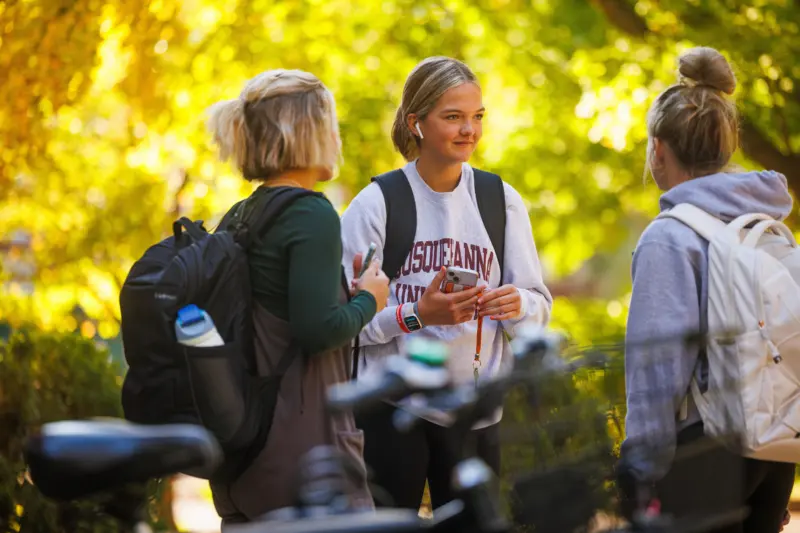 Three young women with backpacks stand outdoors, having a conversation. One holds a phone. They are surrounded by vibrant yellow foliage, and bicycles are visible in the foreground.