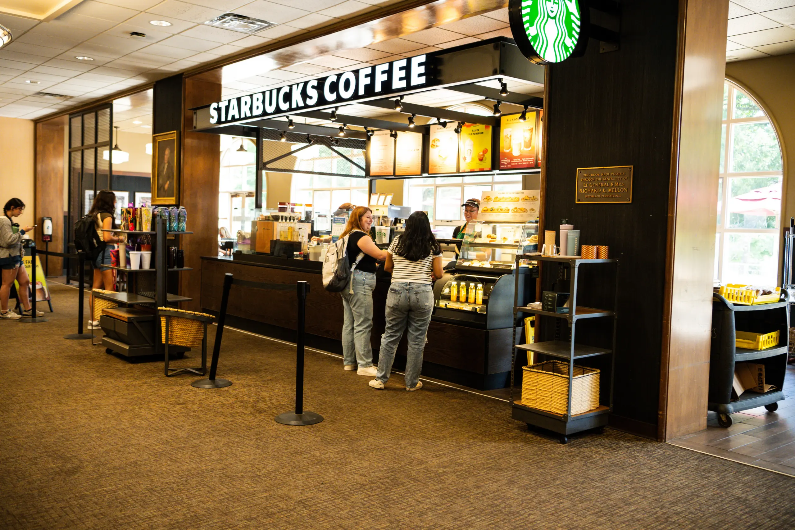 Two women stand at the counter of a Starbucks Coffee inside a building, ordering drinks. The shop, popular for campus life, features a menu above the counter and shelves with products on display. Other customers are nearby.