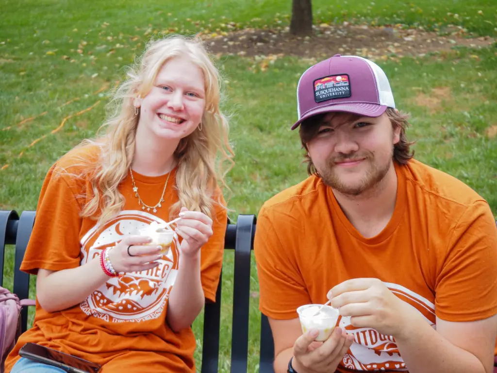 Two people sitting on a bench outdoors, both in bright orange shirts, share smiles and cups of ice cream. About them: the person on the left flaunts long blonde hair, while the one on the right sports a maroon cap. Grass and a tree create a perfect backdrop.