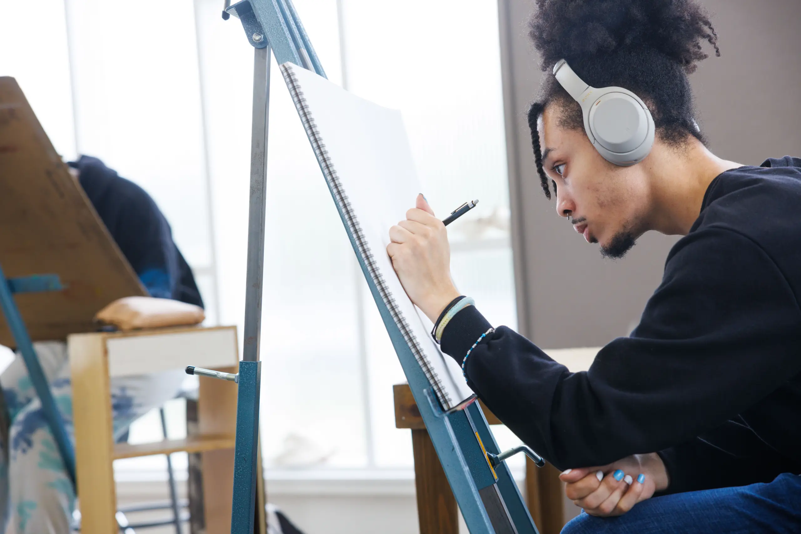 A young person wearing headphones concentrates while sketching on a large notepad at an easel in a bright art studio, considering tuition & cost as they pursue their passion. Another person is blurred in the background, also working at an easel.