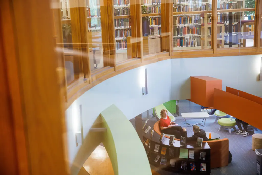 A view through glass of a modern library interior, showing two academics sitting and talking in a curved seating area, surrounded by bookshelves and soft lighting.