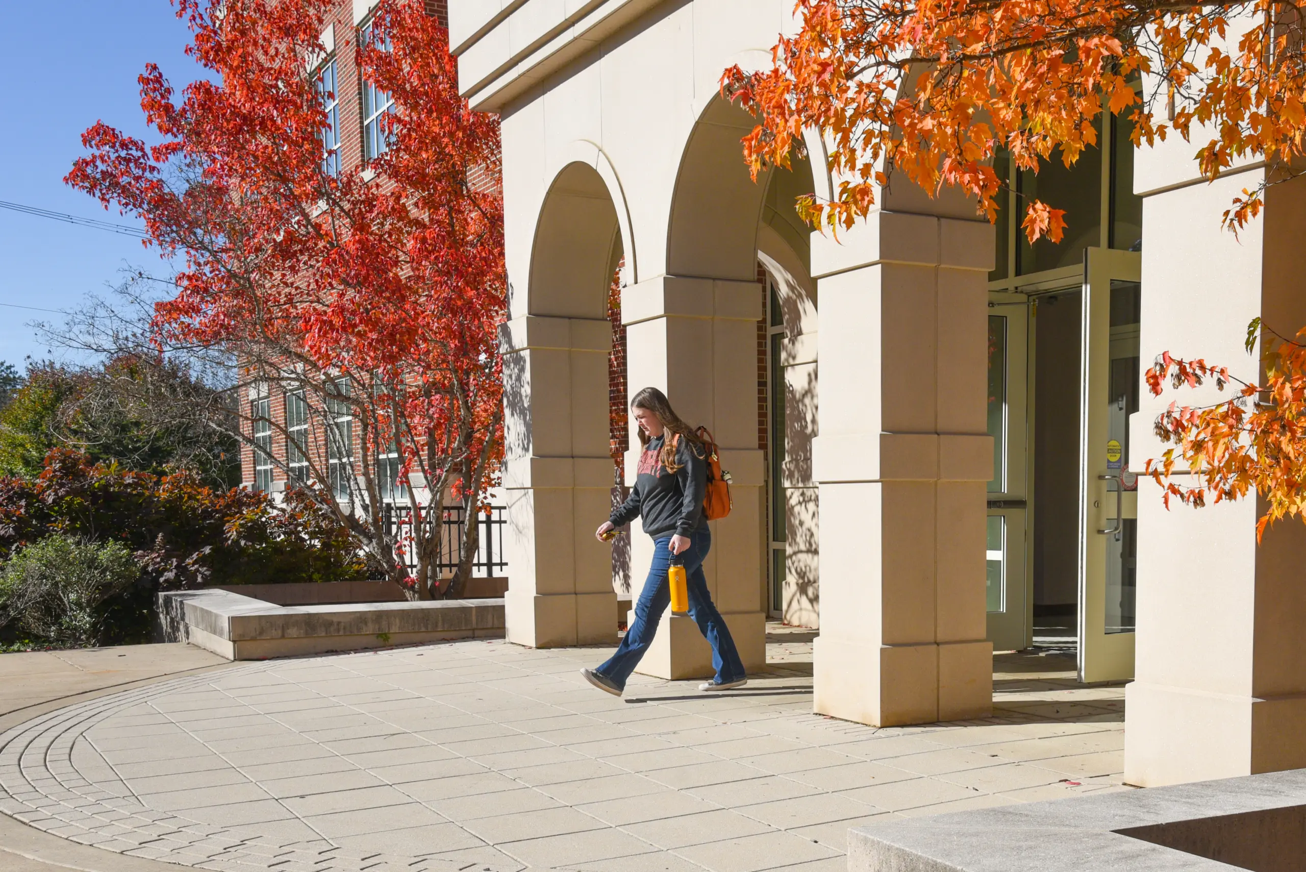 A person walks past a building with arches on a sunny day, surrounded by bright autumn trees. An open door advertising Loans & Financing is visible as the person, carrying a bag and a drink, strolls by.