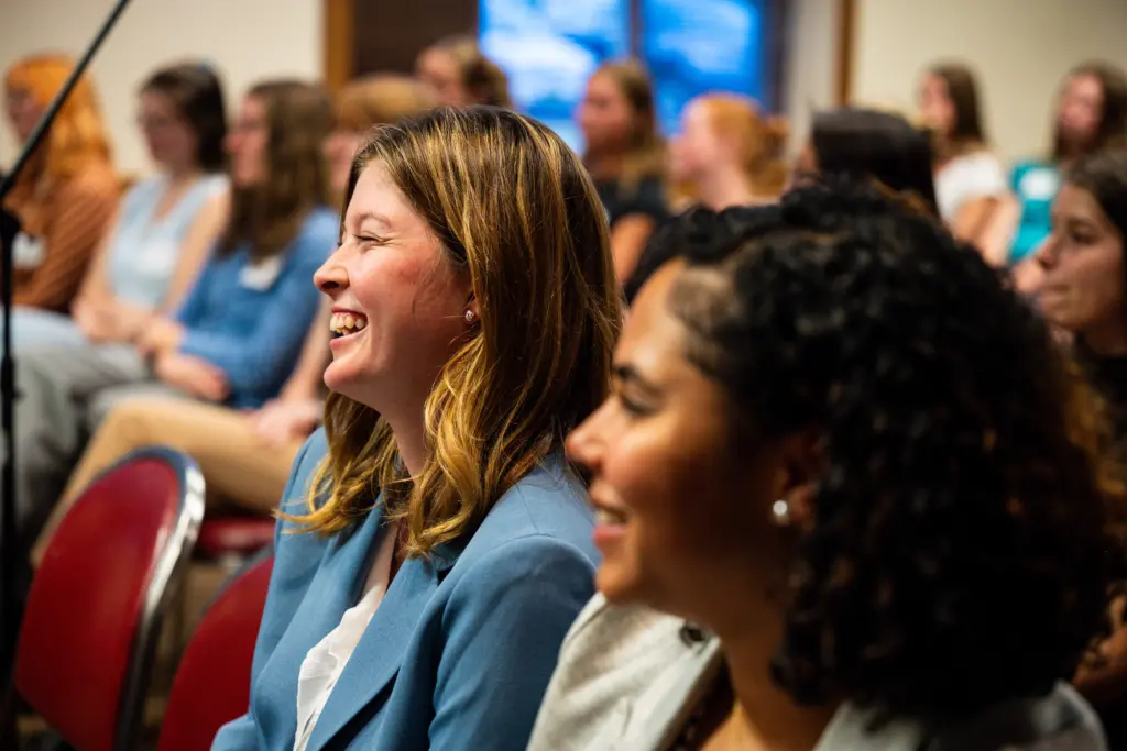 Two women sitting in a crowd are smiling and laughing, while others listen attentively in a brightly lit room during a discussion about tuition & cost.