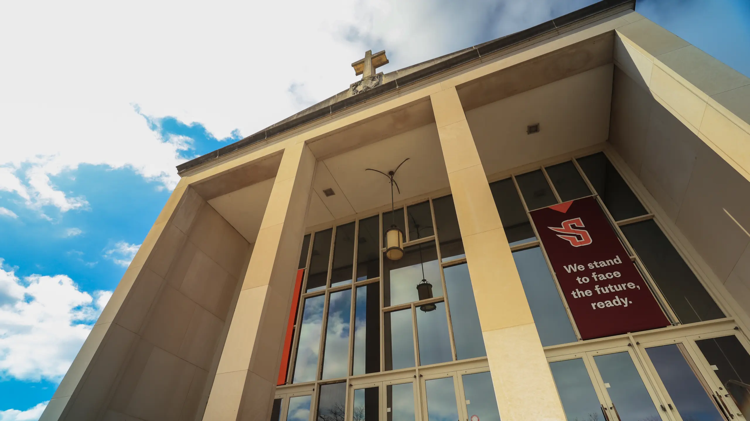 Low-angle view of a building with tall columns, a cross on the roof, large glass windows, and a red banner reading, 