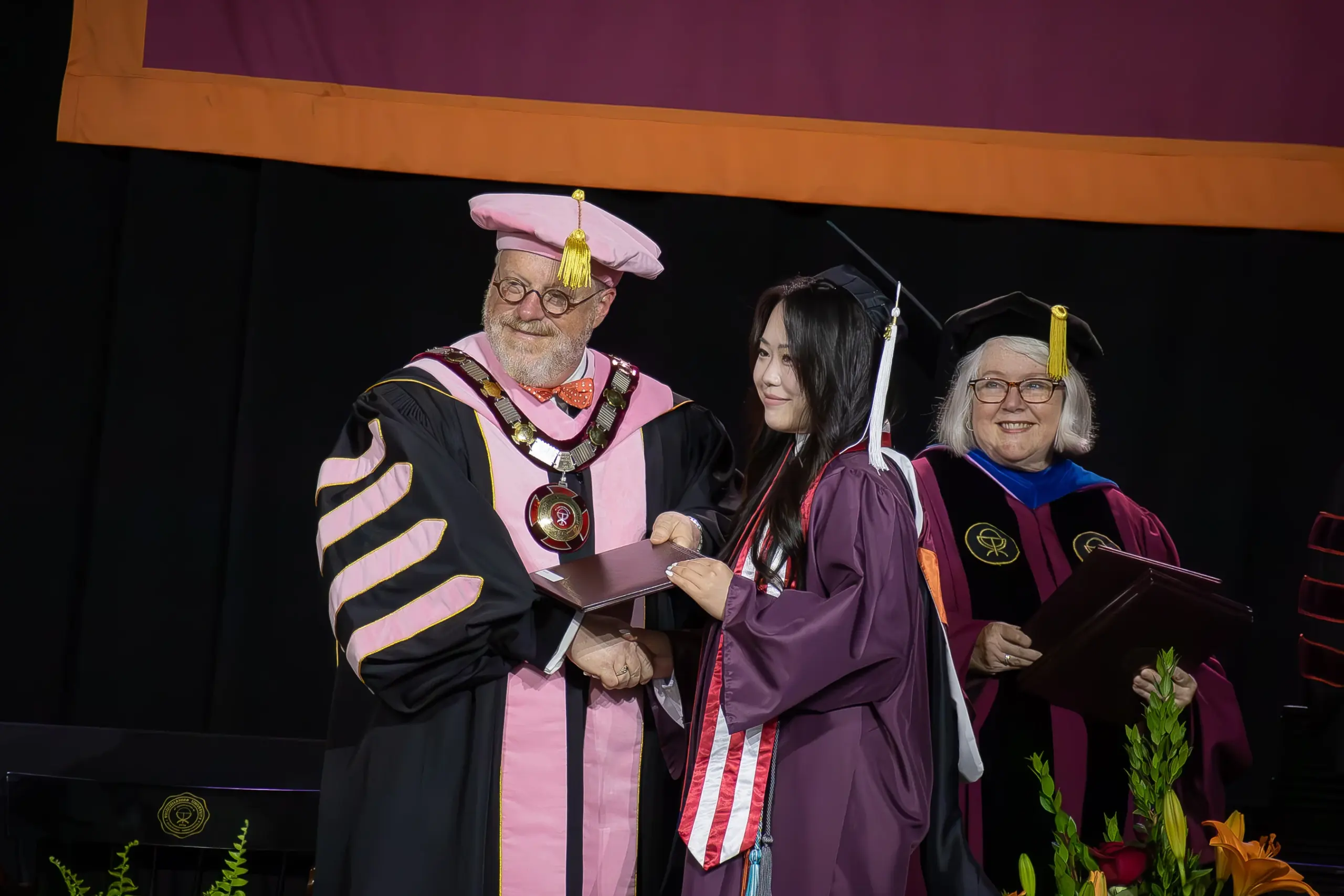 A graduate in a purple cap and gown receives a diploma from a person in pink academic regalia. Another person in black robes stands smiling in the background. The scene is set against a black and maroon backdrop, with flowers visible in front.