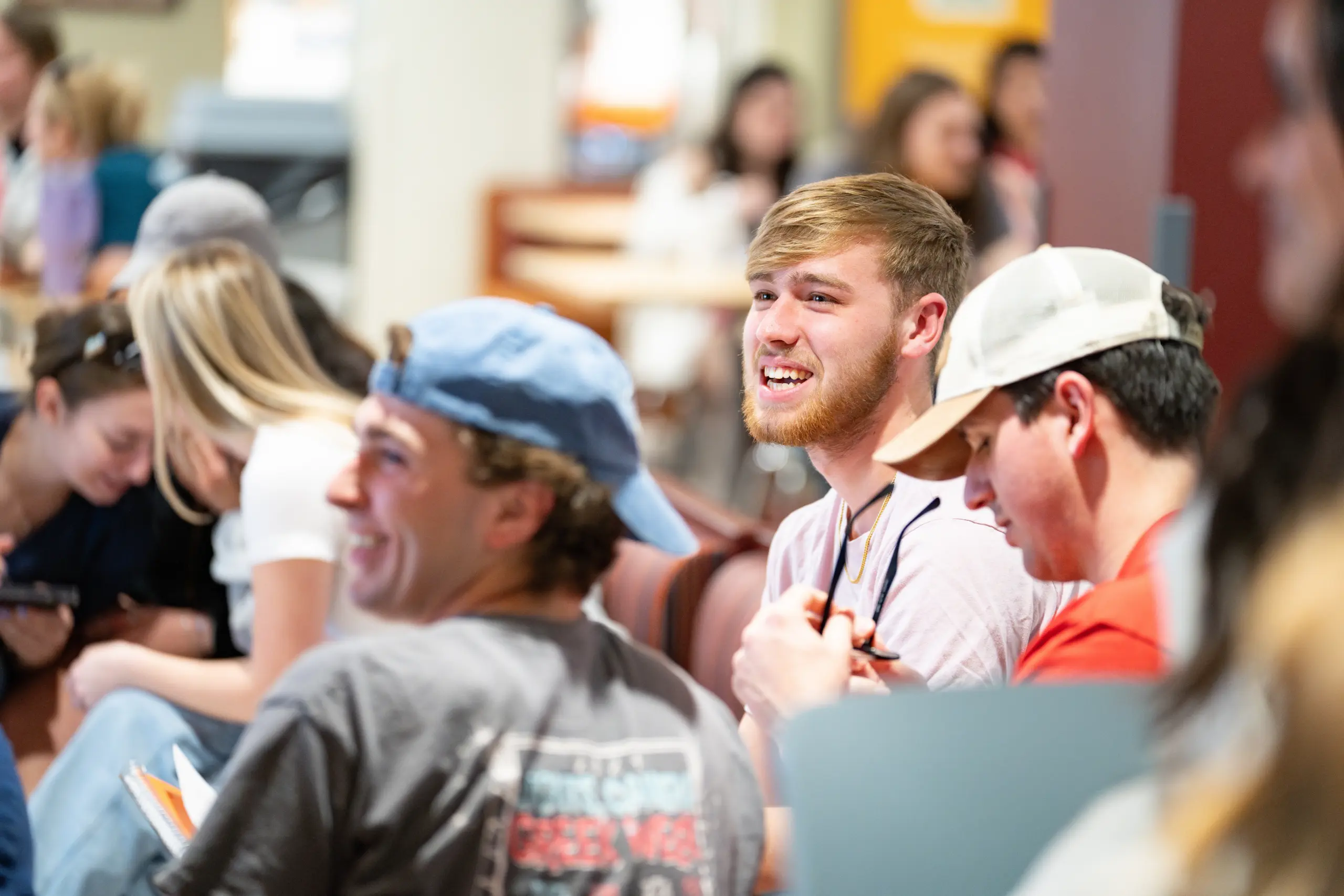 A group of young adults sits together, engaged and smiling in a lively conversation about tuition and financial aid. A bearded man in the center holds sunglasses, appearing cheerful. Others around him wear casual attire, such as caps and T-shirts, in a bright indoor setting.