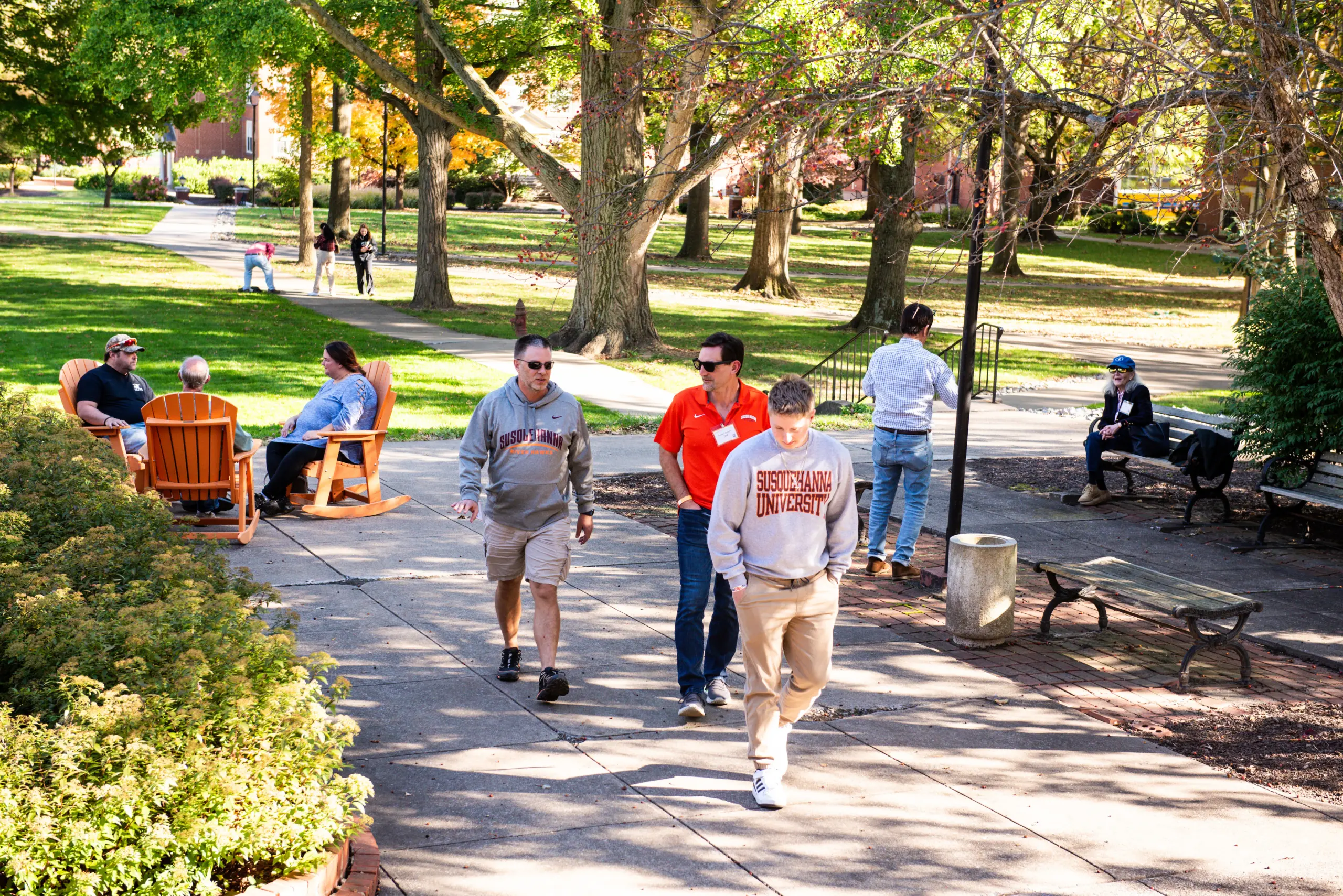 People walking and sitting in a park with benches and trees. Some are wearing casual clothing, while others are in university apparel. The scene is lively with a mix of sun and shade on a paved walkway.