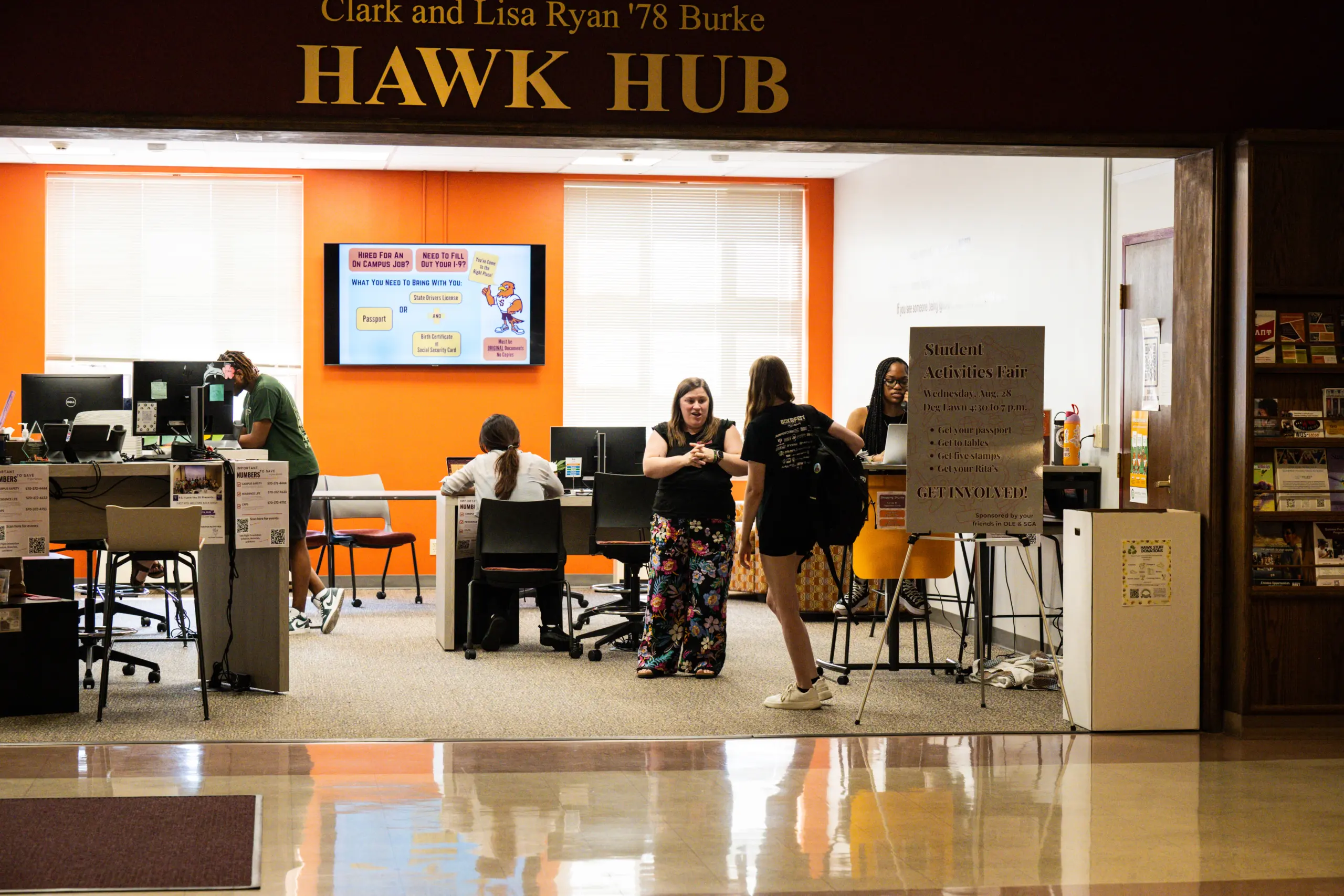 At Hawk Hub, students engage with computers in a vibrant, organized space. A large screen updates them on campus events while a sign promotes an upcoming fair. Desks and chairs are thoughtfully arranged to foster collaboration and productivity.