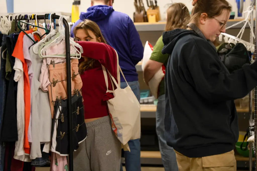 Two people shop at a thrift store, embracing sustainability initiatives. One person browses clothing on a rack, holding a tote bag, while another examines an item nearby. Shelves in the background display various goods, creating a cozy, second-hand shopping atmosphere.