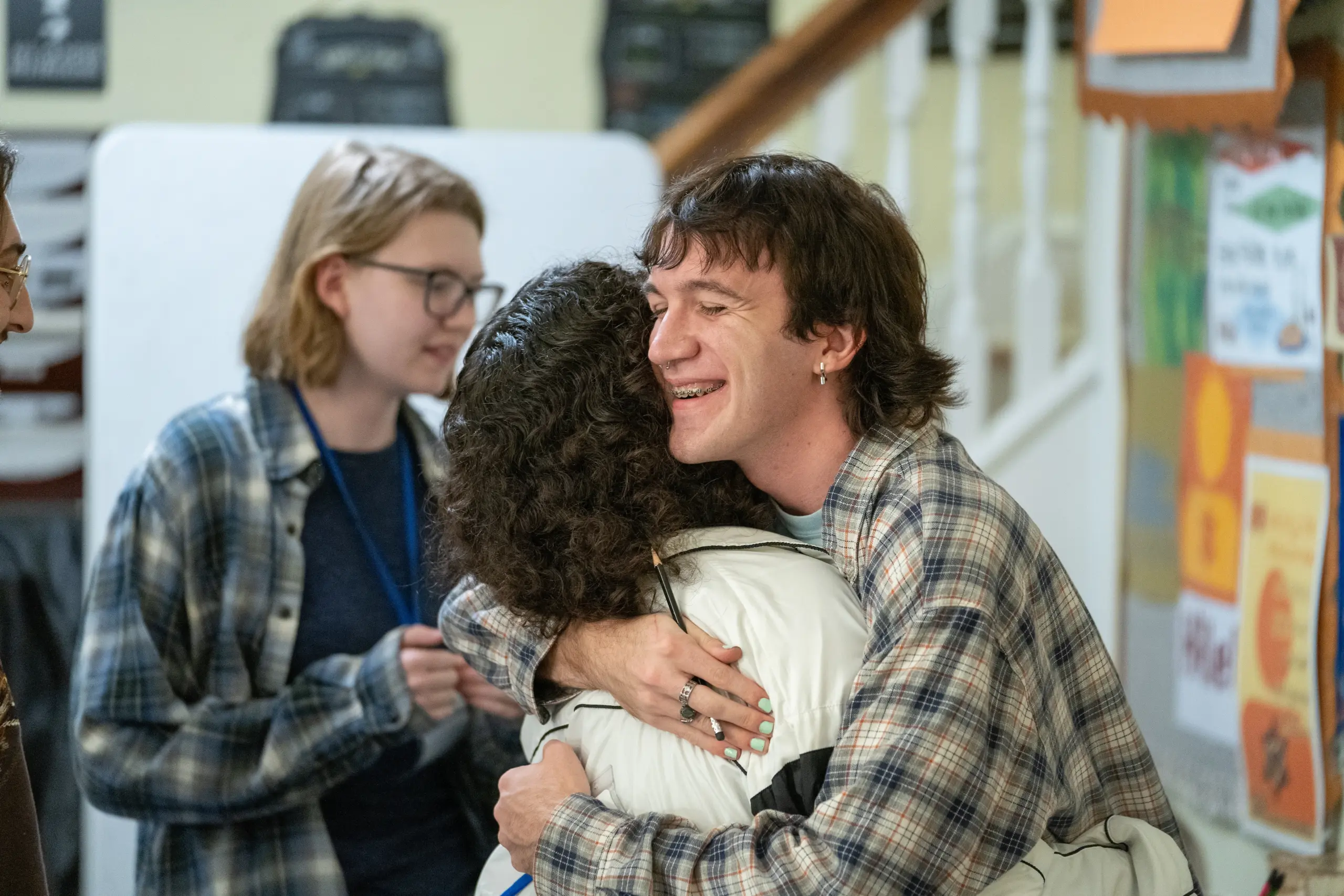 A young man in a plaid shirt warmly hugs a woman with curly hair indoors, capturing the spirit of campus life, while another person with glasses and a blue lanyard stands nearby, smiling. Colorful posters are visible in the background.