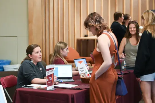 A woman in an orange dress approaches a registration table staffed by Student Services, where two women sit behind laptops. The table is adorned with signs and a basket. Nearby, people stand in a room with wooden walls.