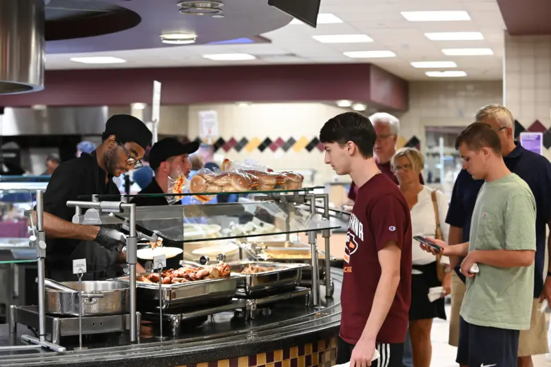 A bustling cafeteria scene unfolds as people line up for food. Amidst the chatter, a server donning gloves and a hat places meals on trays. Customers, some holding eco-friendly trays or phones, await their turn, while the decor subtly emphasizes sustainability initiatives in dining.