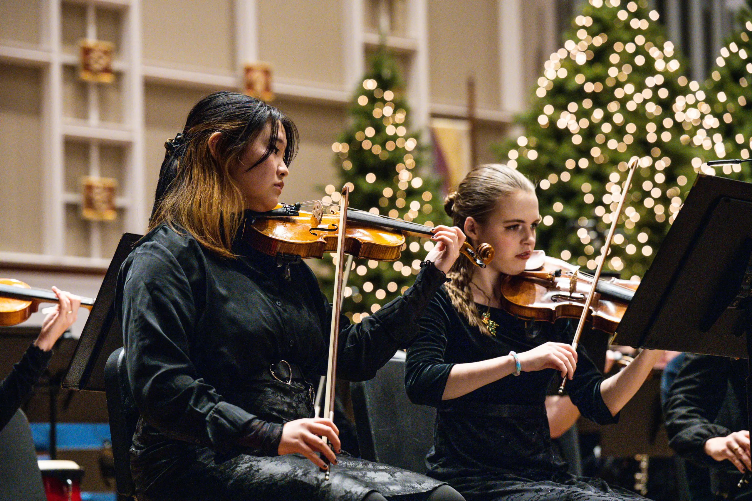 Two women dressed in black play violins in an orchestra, with decorated Christmas trees and festive lights in the background.