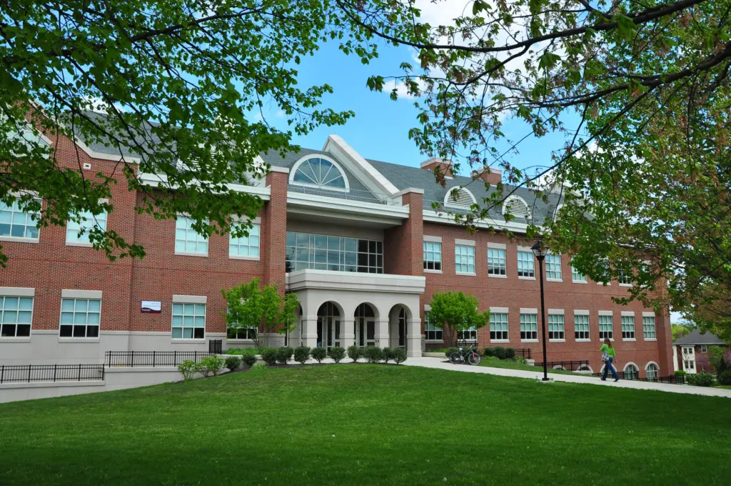 A large, two-story brick building with white trim and arched windows sits on a lush green lawn, embodying sustainability. Trees with fresh leaves frame the scene as two people stroll along a path in front of the building under a vibrant blue sky.
