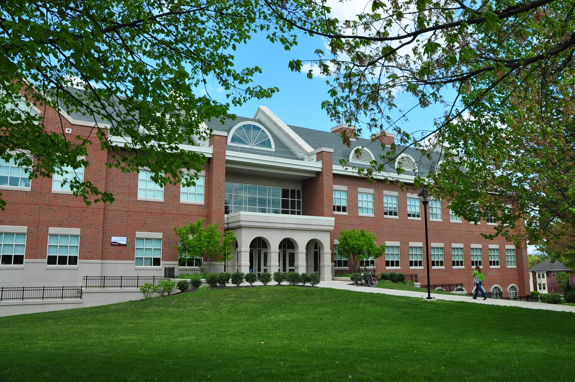 A large, two-story brick building with white trim and arched windows sits on a lush green lawn, embodying sustainability. Trees with fresh leaves frame the scene as two people stroll along a path in front of the building under a vibrant blue sky.