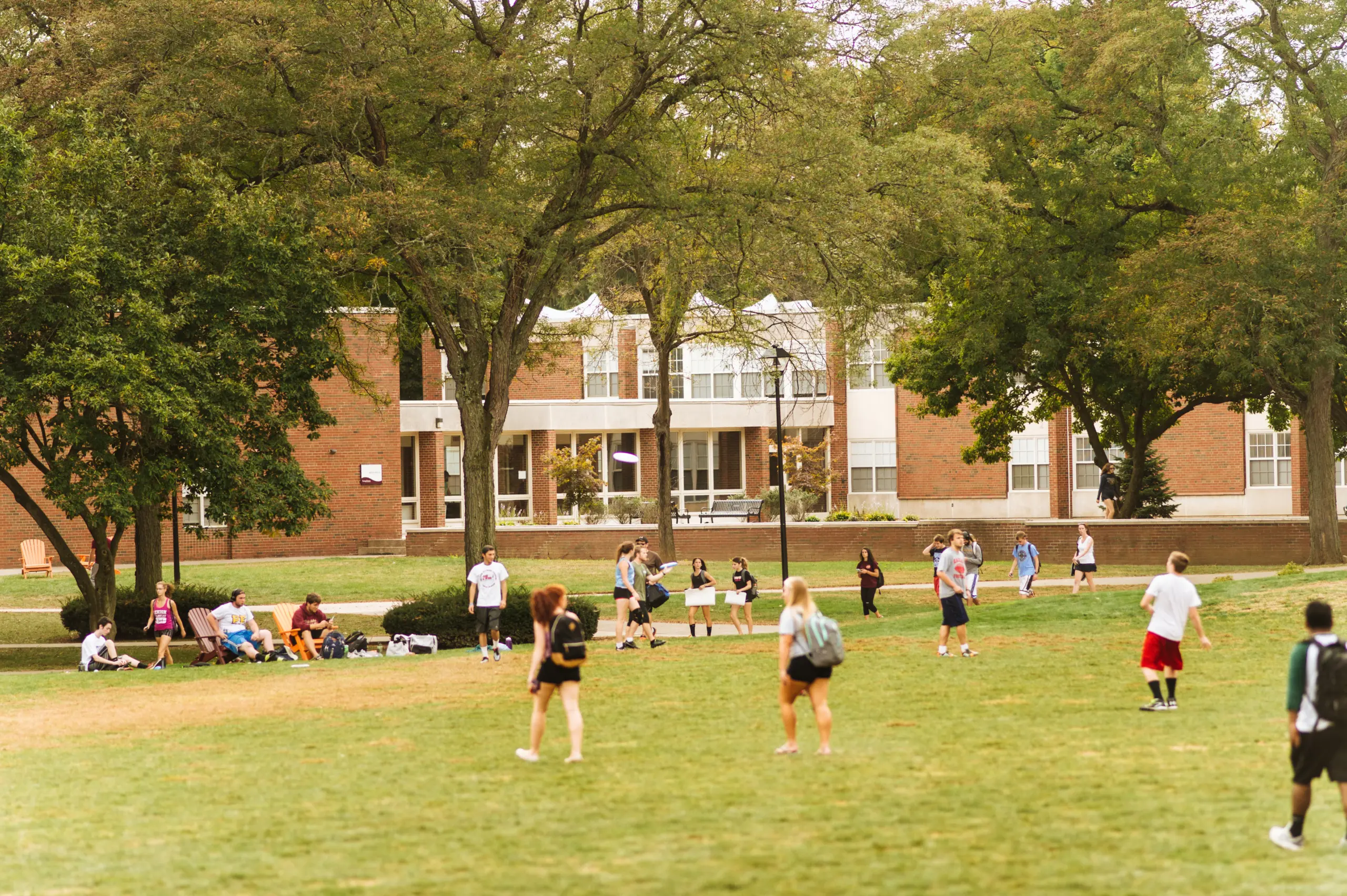 People are playing sports on a grassy field surrounded by trees. A brick building is in the background. Some individuals are standing and observing, while others are actively participating in a game or walking across the field.