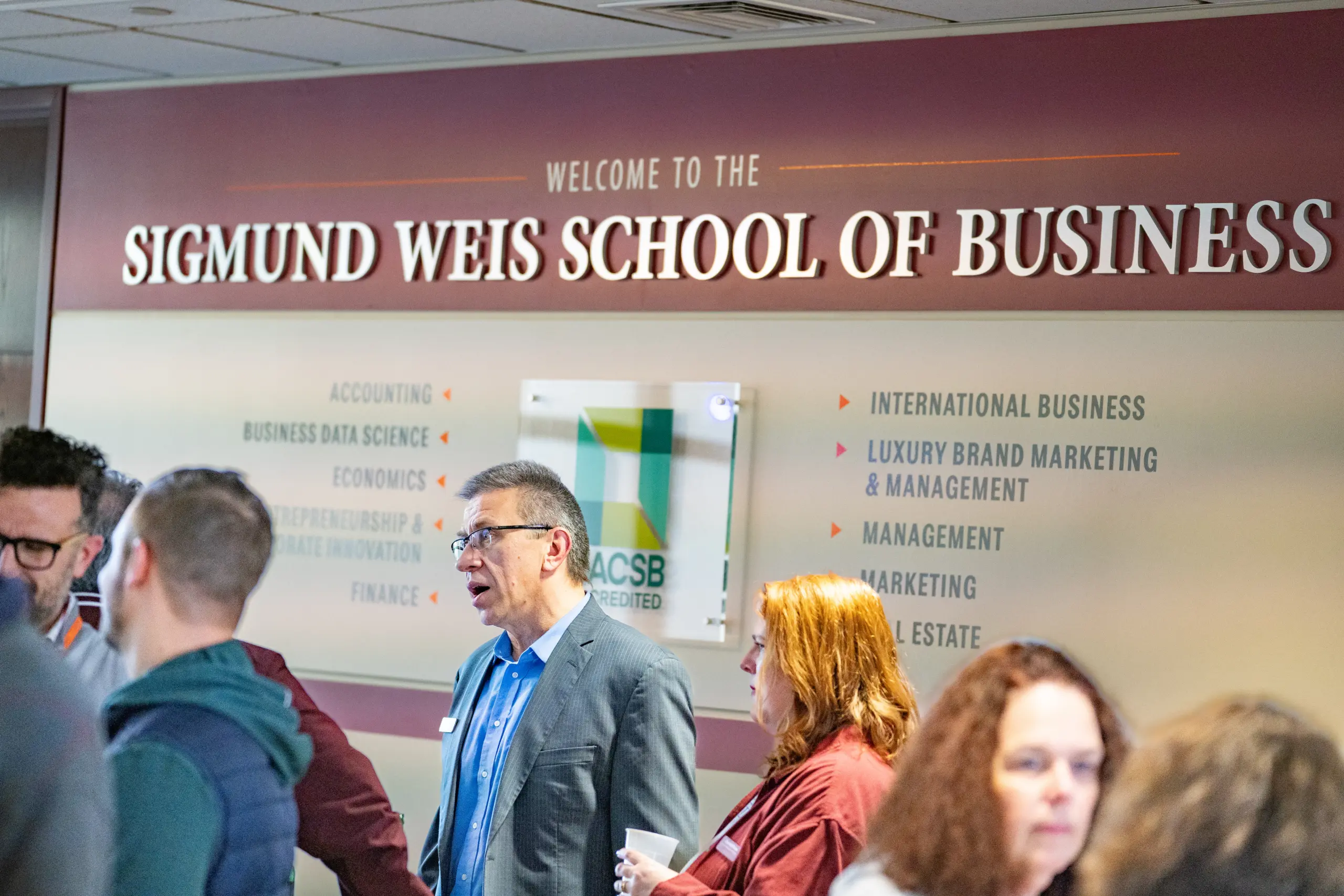 People standing in a hallway of the Sigmund Weis School of Business. The wall displays the school's name and a list of subjects, such as accounting and international business. A sign shows accreditation from AACSB.
