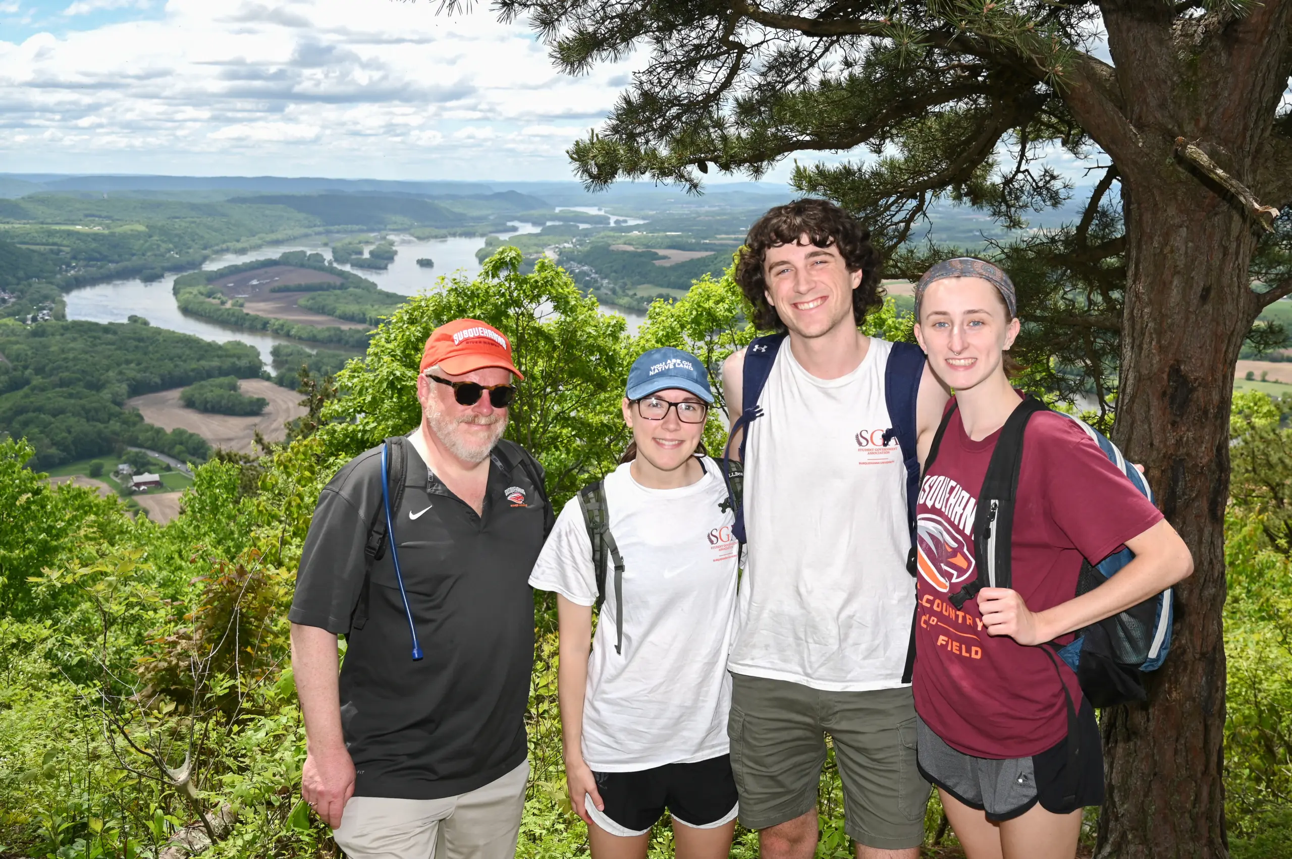 About four friends strike a pose together on a scenic overlook, surrounded by green trees. Behind them, a winding river and rolling hills stretch into the distance under a partly cloudy sky. The group is dressed in casual hiking attire.