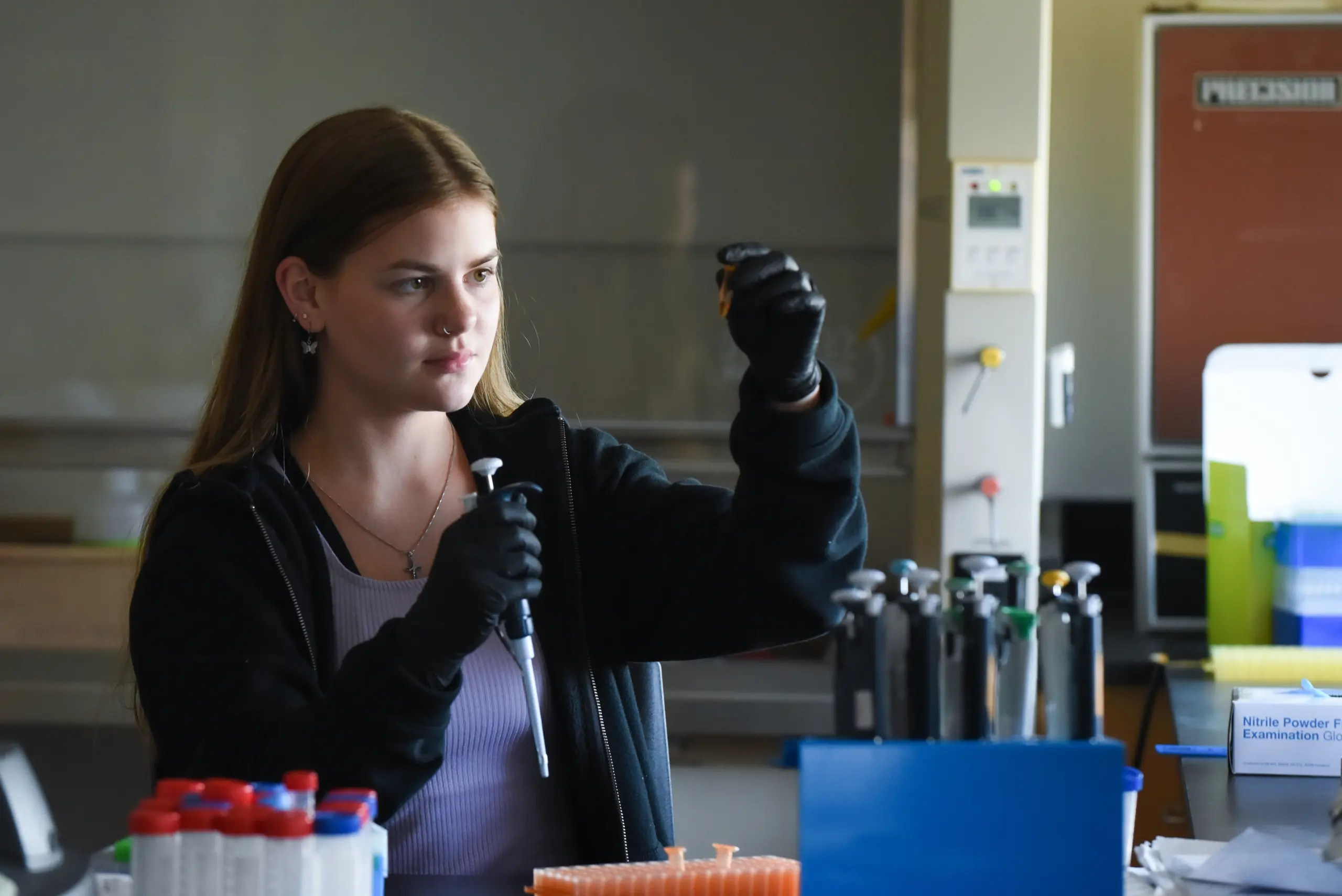 A woman in a black jacket and gloves is using lab equipment to inspect a sample in a laboratory. She is focused on her work, surrounded by test tubes and pipettes on a desk.