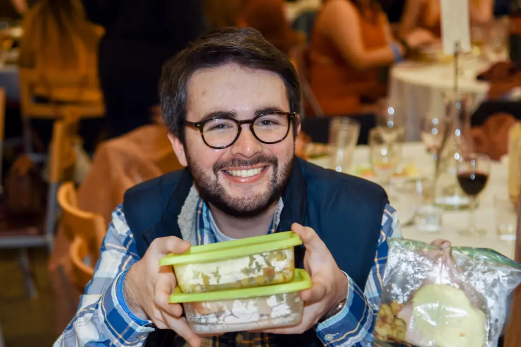 A man with glasses and a beard smiles at the camera, showcasing his commitment to sustainability initiatives by holding two eco-friendly food containers with green lids. He's dressed in a plaid shirt and dark vest, while behind him, people sit at tables adorned with plates and glasses.