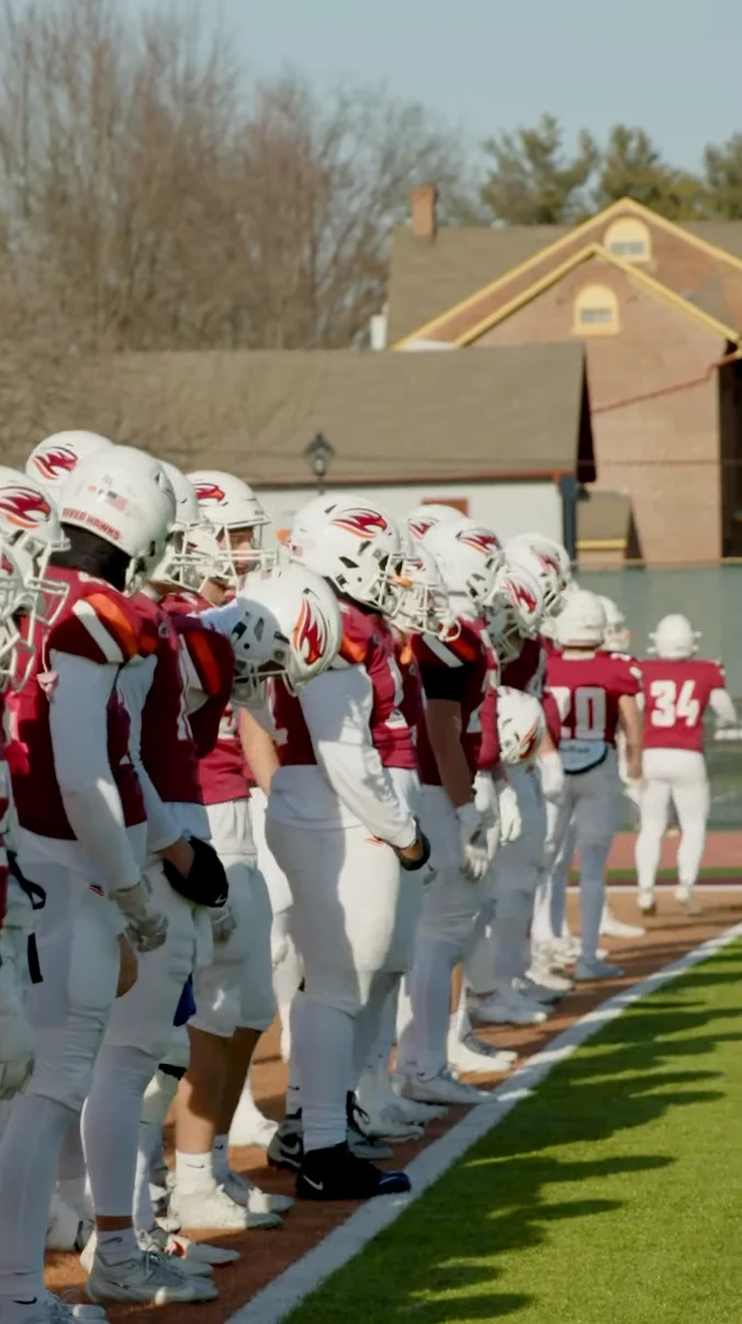 A row of American football players in red and white uniforms and helmets stand on the sidelines, watching the game—a classic scene of campus life with houses and trees visible in the background.