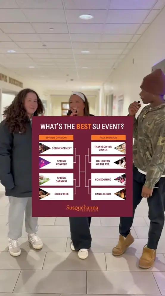 Three students stand in a hallway while one holds a microphone. In front of them is a bracket chart titled 