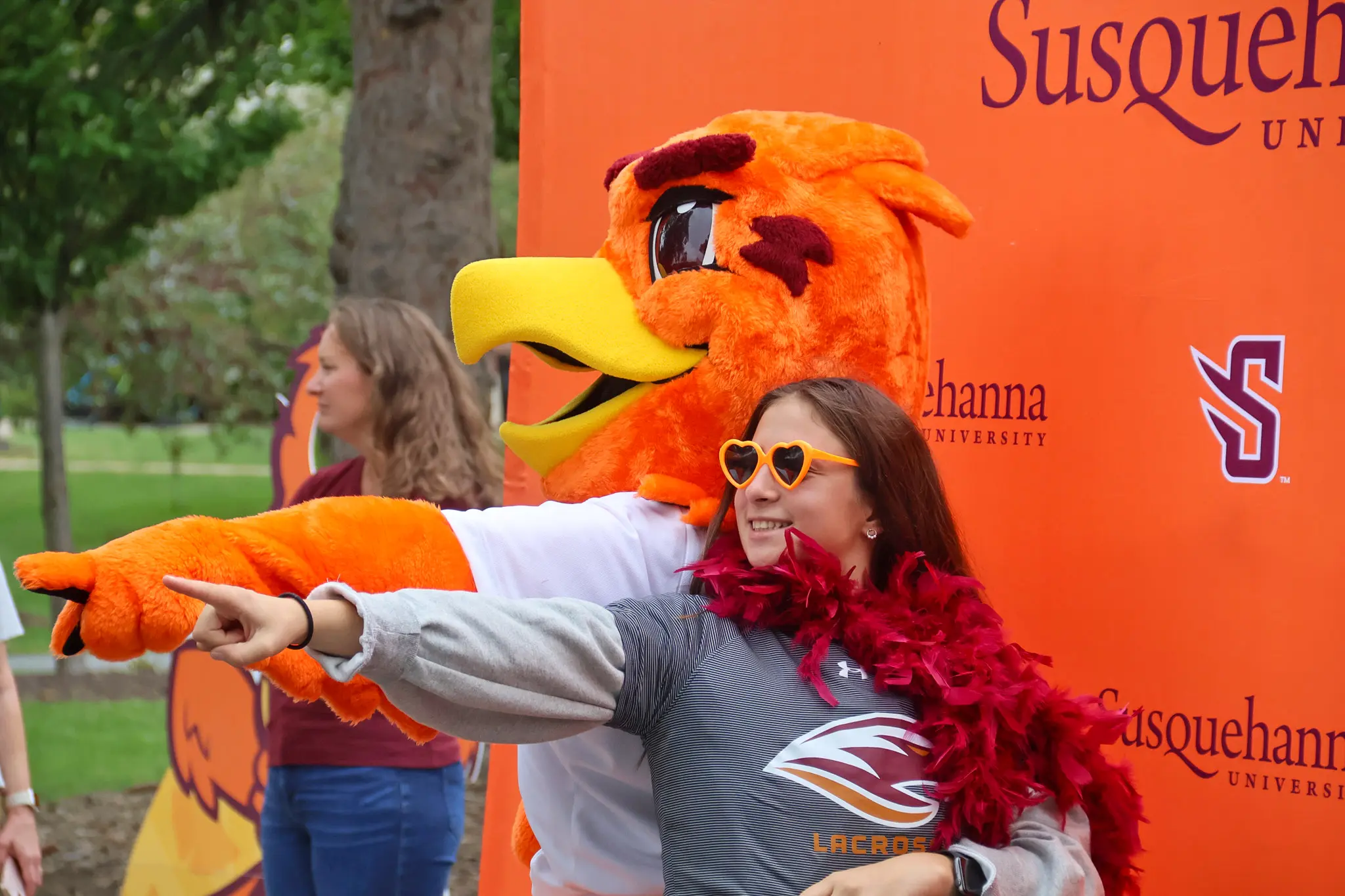 About: A person wearing heart-shaped sunglasses and a feather boa poses with an orange bird mascot; both are smiling, pointing, and having fun in front of a 体育买球官网 University backdrop.