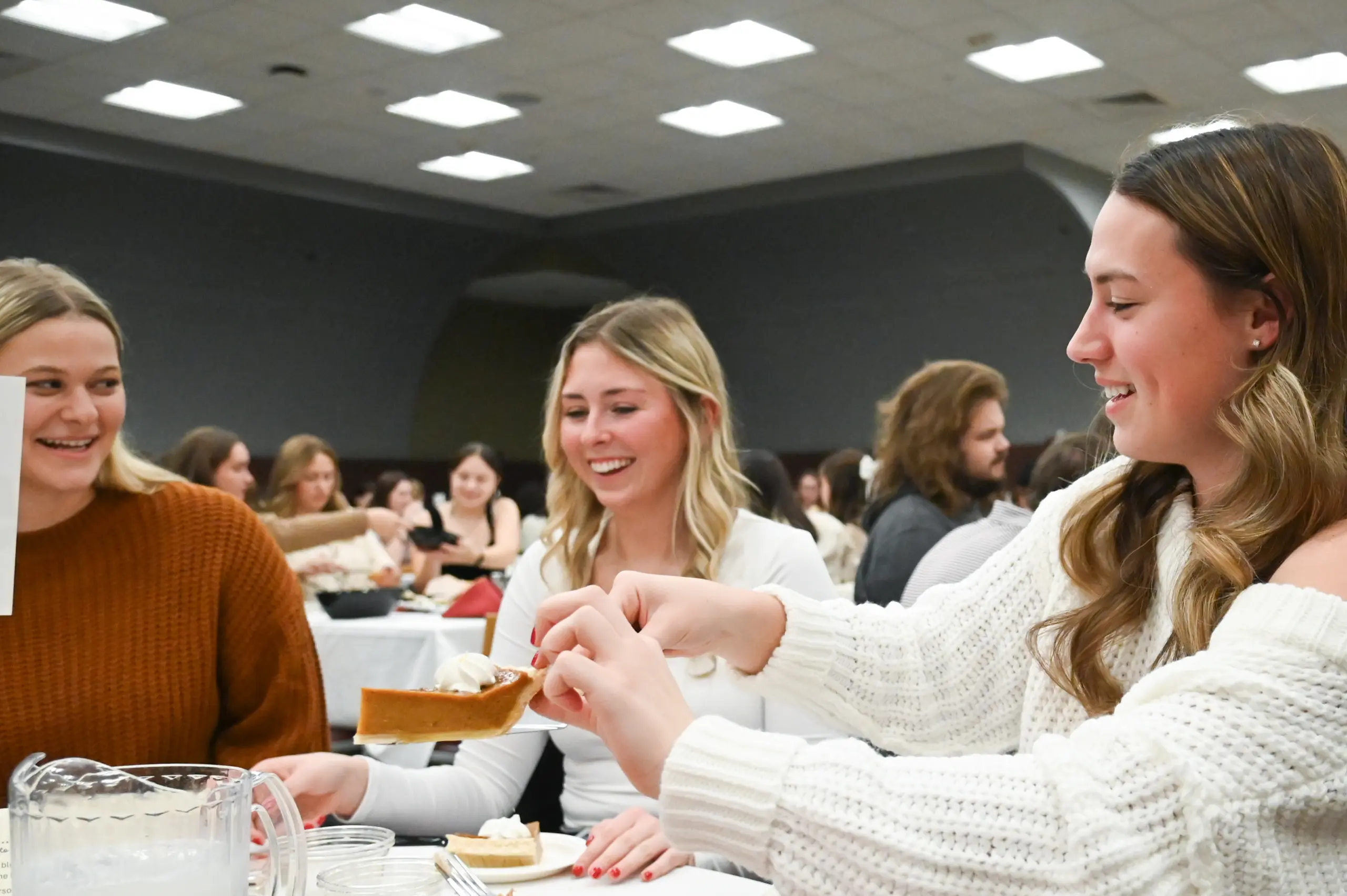About three young women sitting at a round table, smiling and enjoying dessert. One woman in a white sweater serves a slice of pie as the room buzzes with people seated at similar tables under bright lighting.