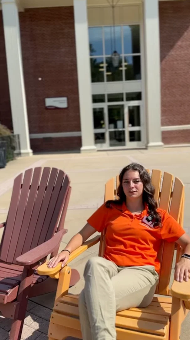 A person in an orange polo shirt and khaki pants sits relaxed on a wooden outdoor chair in front of a building with large glass doors and brick walls.