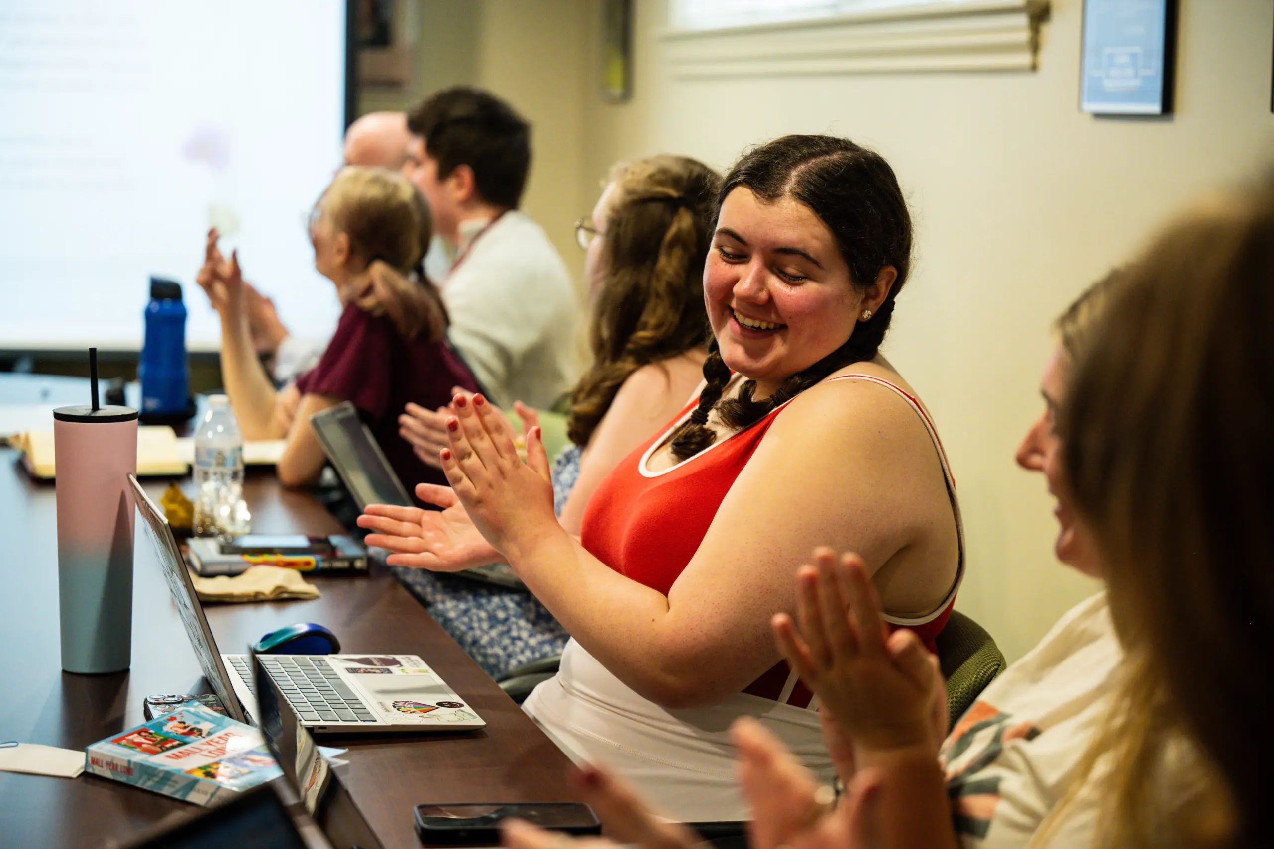 A group of people sitting at a table in a meeting room, applauding. Some have laptops and water bottles in front of them. The focus is on a smiling woman in a red and white outfit, clapping her hands.