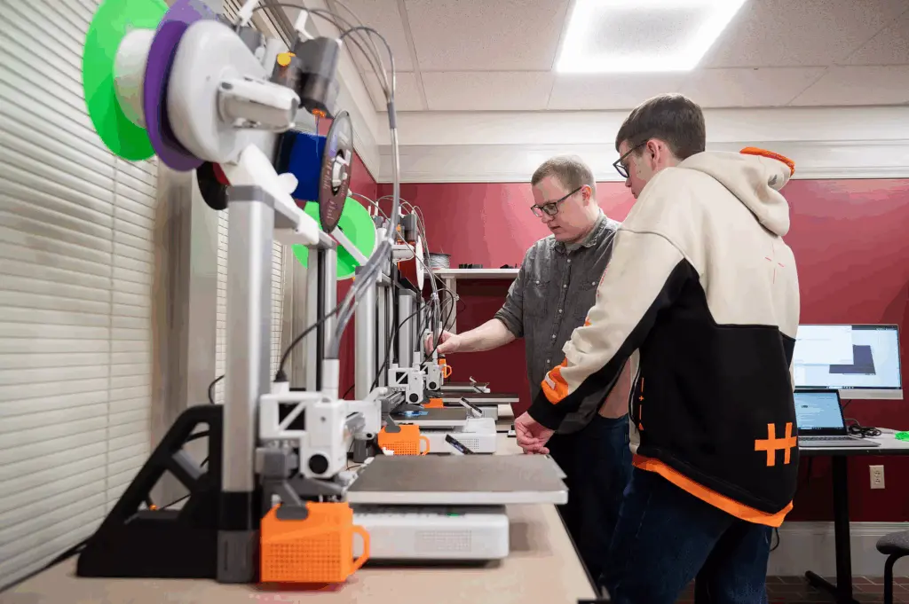 Two people stand in front of 3D printers on a table, examining one of the machines in a well-lit room with red walls, computers, and modern Building and Spaces elements in the background.