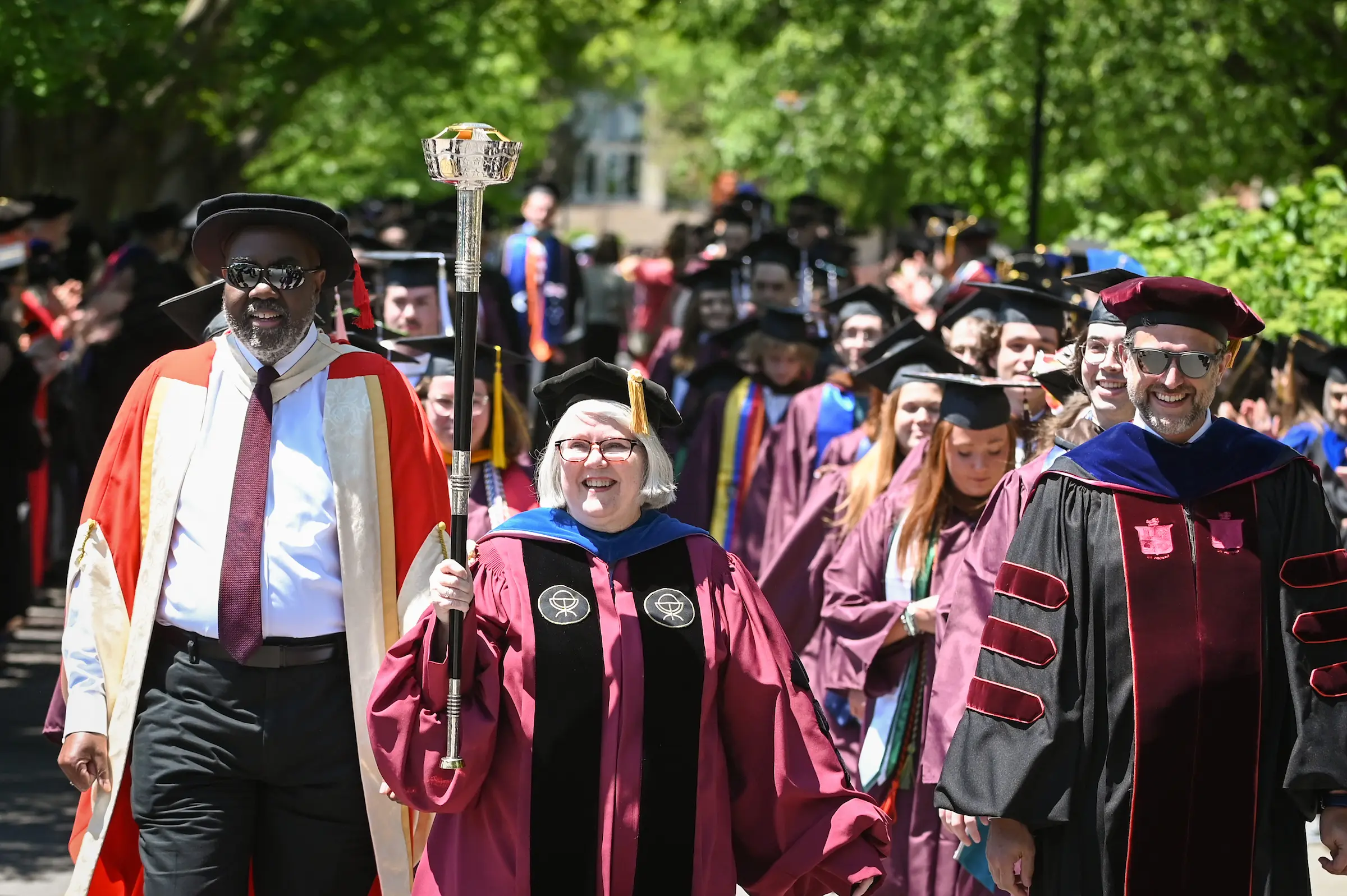 A group of faculty members in academic regalia lead a graduation procession outdoors, with one person holding a ceremonial mace and graduates following behind on a sunny day.