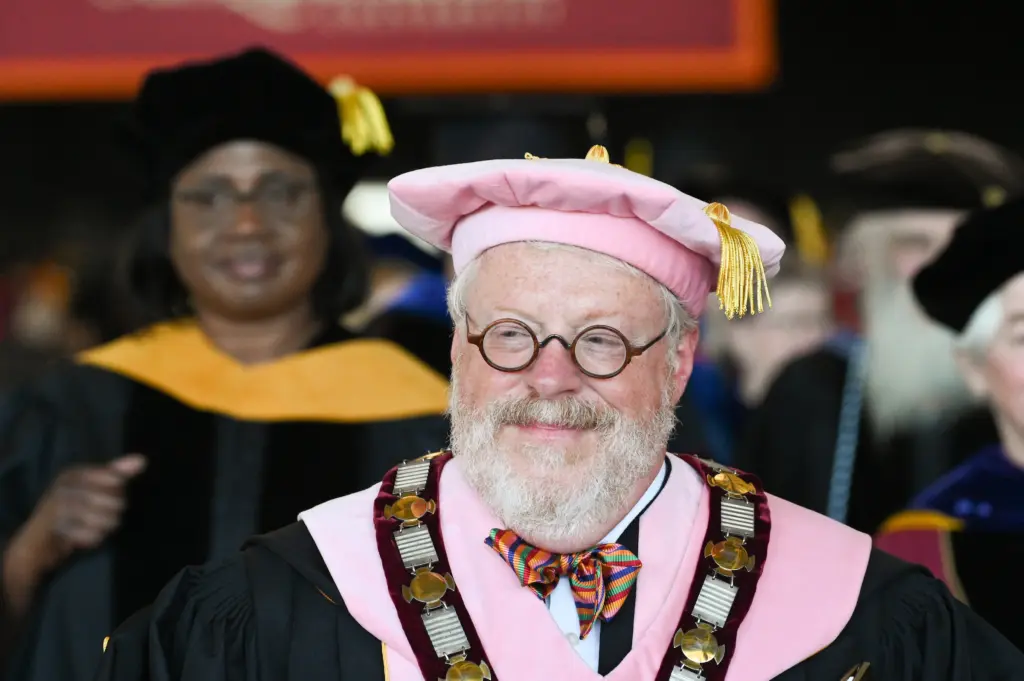A smiling man, representing our leadership, wears academic regalia, a pink cap, and a colorful bow tie at a graduation ceremony. Others in similar attire are blurred in the background.