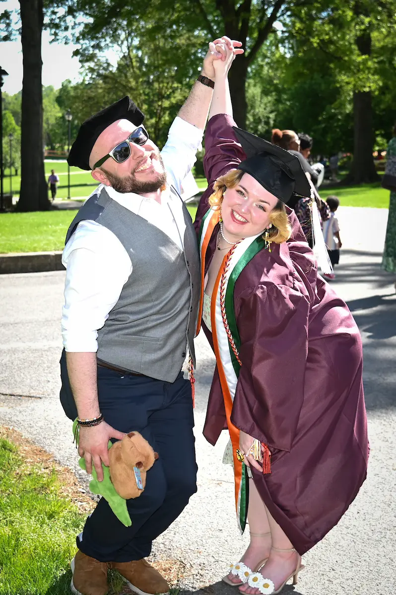 A smiling graduate in a maroon gown and cap celebrates Commencement joyfully with a man in sunglasses and a beret, both holding hands and raising their arms while standing outside on a sunny day.