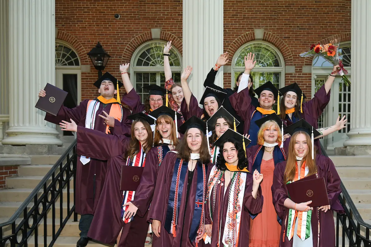 A group of graduates in caps and gowns cheer and pose together on steps in front of a brick building with columns, holding diplomas and flowers, celebrating their Commencement achievement.