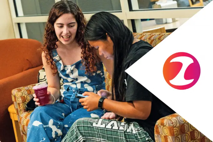 Two young women sit on a patterned couch. One holds a cup and smiles while the other looks at her phone, possibly checking an admission update. A large stylized logo in pink and orange appears on the right side of the image.