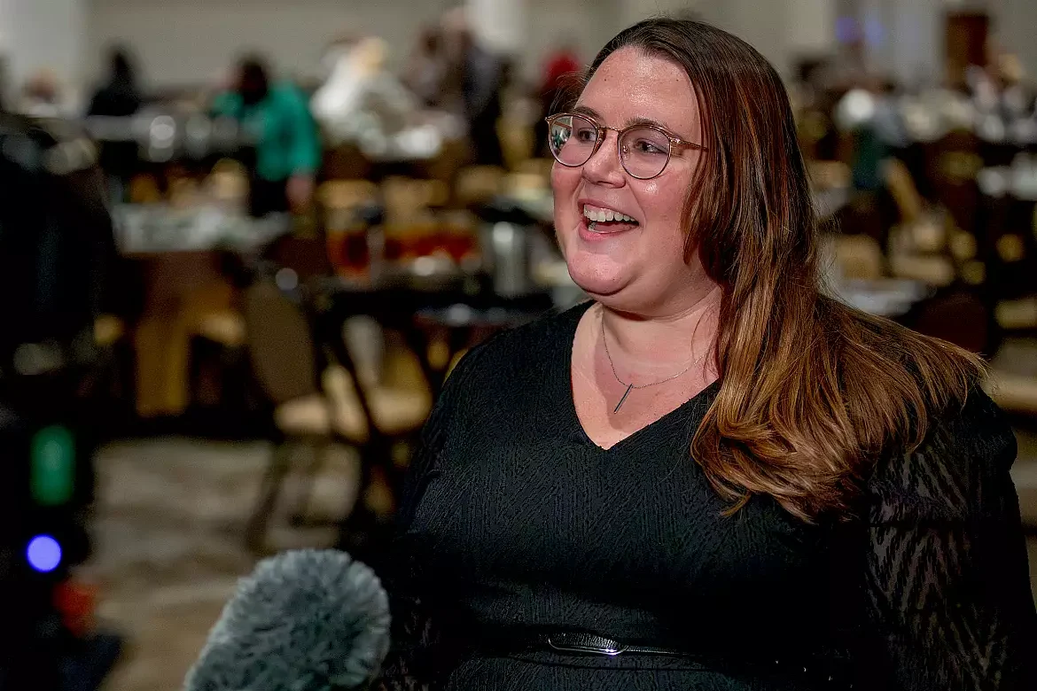 A woman with long brown hair and glasses, wearing a black top, smiles while being interviewed with a microphone in front of her; people and tables are visible in the blurred background.