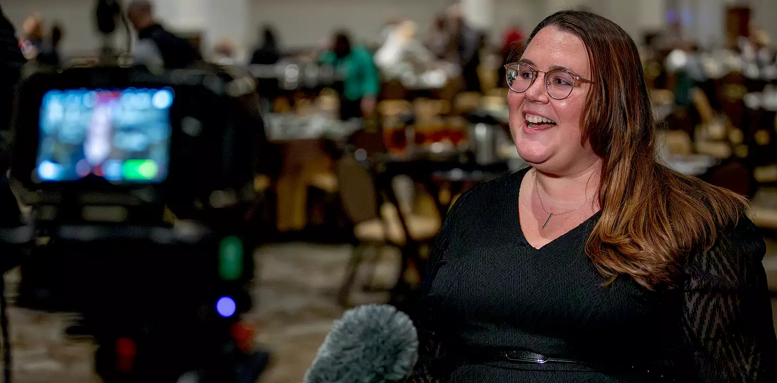 A woman with long brown hair and glasses, wearing a black top, smiles while being interviewed with a microphone in front of her; people and tables are visible in the blurred background.